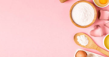 Wooden baking tools and ingredients, including eggs and flour, arranged on a pastel pink background.