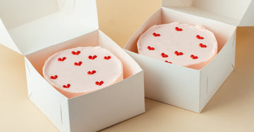 Two round cakes with pink frosting and red hearts on top. The cakes are in white boxes, ready for transport.